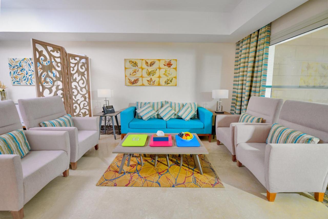 Tolip Resort El Galala Majestic Ain Sukhna Bagian luar foto The photo shows a stylish and colorful living room. It features a bright blue sofa with patterned cushions, complemented by two light gray armchairs. In front of the sofa, there's a wooden coffee table with vibrant trays in pink, green, and yellow. A