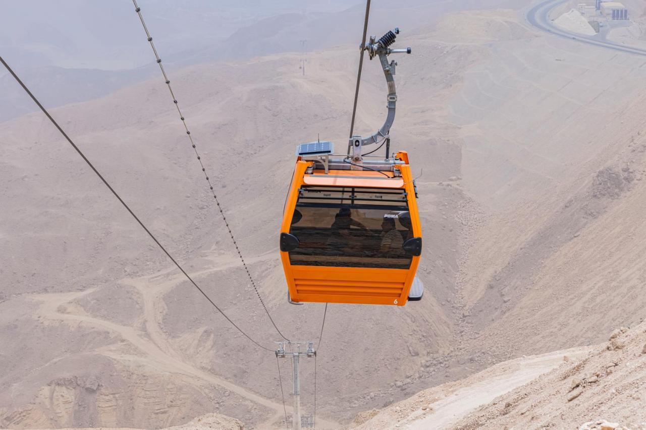 Tolip Resort El Galala Majestic Ain Sukhna Bagian luar foto The photo shows an orange cable car suspended in the air, traveling along a set of cables. It appears to be located in a mountainous or hilly area, with rugged terrain and dry, rocky landscapes visible below. The background features slopes that seem 