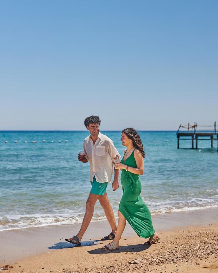 Tolip Resort El Galala Majestic Ain Sukhna Bagian luar foto The photo shows a couple walking along a beach. The man is wearing a white short-sleeved shirt and green shorts, while the woman is dressed in a flowing green dress. They both appear to be enjoying the pleasant weather, with the ocean in the backgrou