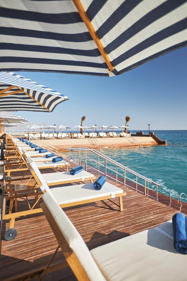Tolip Resort El Galala Majestic Ain Sukhna Bagian luar foto The photo depicts a serene outdoor area by the waterfront. There are several lounge chairs arranged in a row, each equipped with blue towels. Large, striped umbrellas provide shade over some of the chairs. In the background, there is a wooden deck th