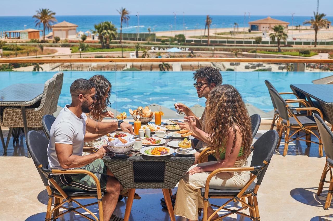 Tolip Resort El Galala Majestic Ain Sukhna Bagian luar foto The photo shows a group of five people enjoying a meal together outdoors, likely at a restaurant or cafe. They are seated at a table with a beautiful view of a swimming pool and the sea in the background. The table is filled with various dishes, and 