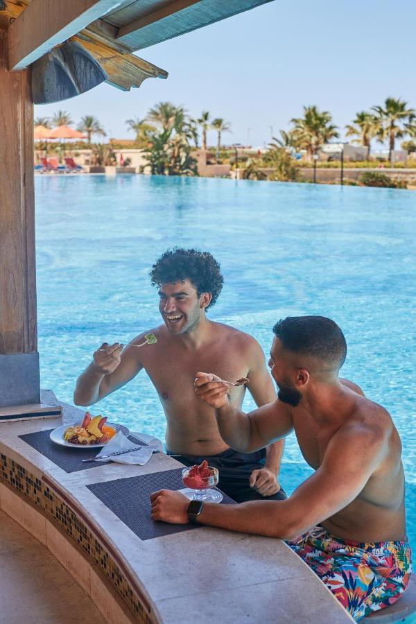 Tolip Resort El Galala Majestic Ain Sukhna Bagian luar foto The photo features two young men enjoying a meal together. They are sitting at a poolside table with a clear blue swimming pool in the background. The men are shirtless, suggesting a casual, relaxed atmosphere, likely in a warm environment. One man i