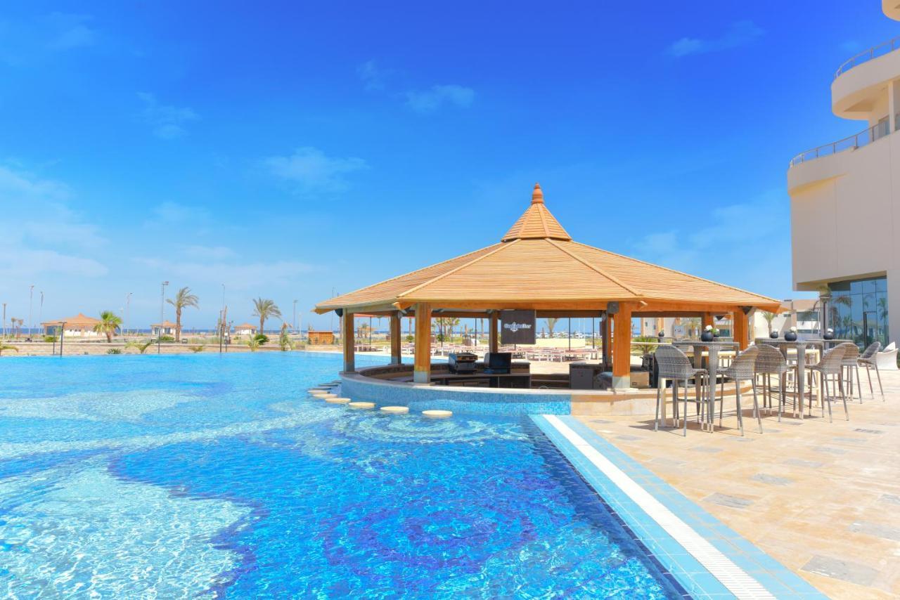 Tolip Resort El Galala Majestic Ain Sukhna Bagian luar foto The photo shows a beautiful outdoor setting featuring a large swimming pool with clear blue water. In the center, there is a thatched-roof gazebo, which serves as a shaded seating area. Surrounding the gazebo are lounge chairs and a bar area with hig