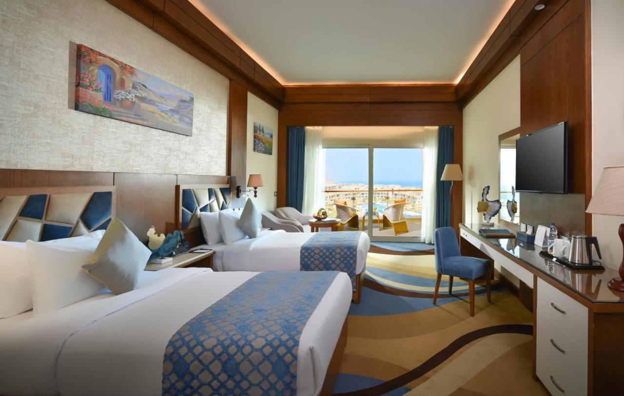 Tolip Resort El Galala Majestic Ain Sukhna Bagian luar foto The photo shows a well-decorated hotel room featuring two beds with decorative pillows. The room has wooden accents and modern furnishings, including a desk, a television, and lamps. Large windows flood the space with natural light, offering a view o