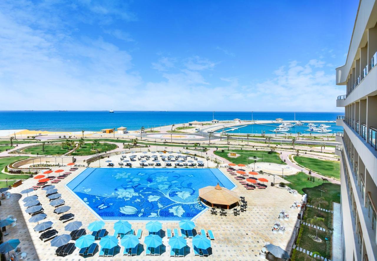 Tolip Resort El Galala Majestic Ain Sukhna Bagian luar foto The photo shows a beautiful outdoor view of a resort area featuring a large swimming pool with a decorative underwater design. Surrounding the pool are numerous sun loungers and umbrellas in a variety of colors. In the background, there is a scenic c