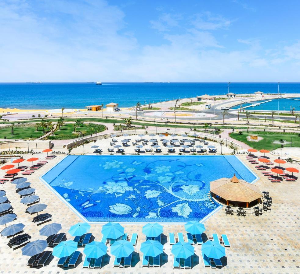Tolip Resort El Galala Majestic Ain Sukhna Bagian luar foto The photo features a large swimming pool with a decorative design on its surface, surrounded by sun loungers and umbrellas. In the background, there is a view of the sea, with a clear blue sky and a few scattered clouds. The area is well-maintained, 