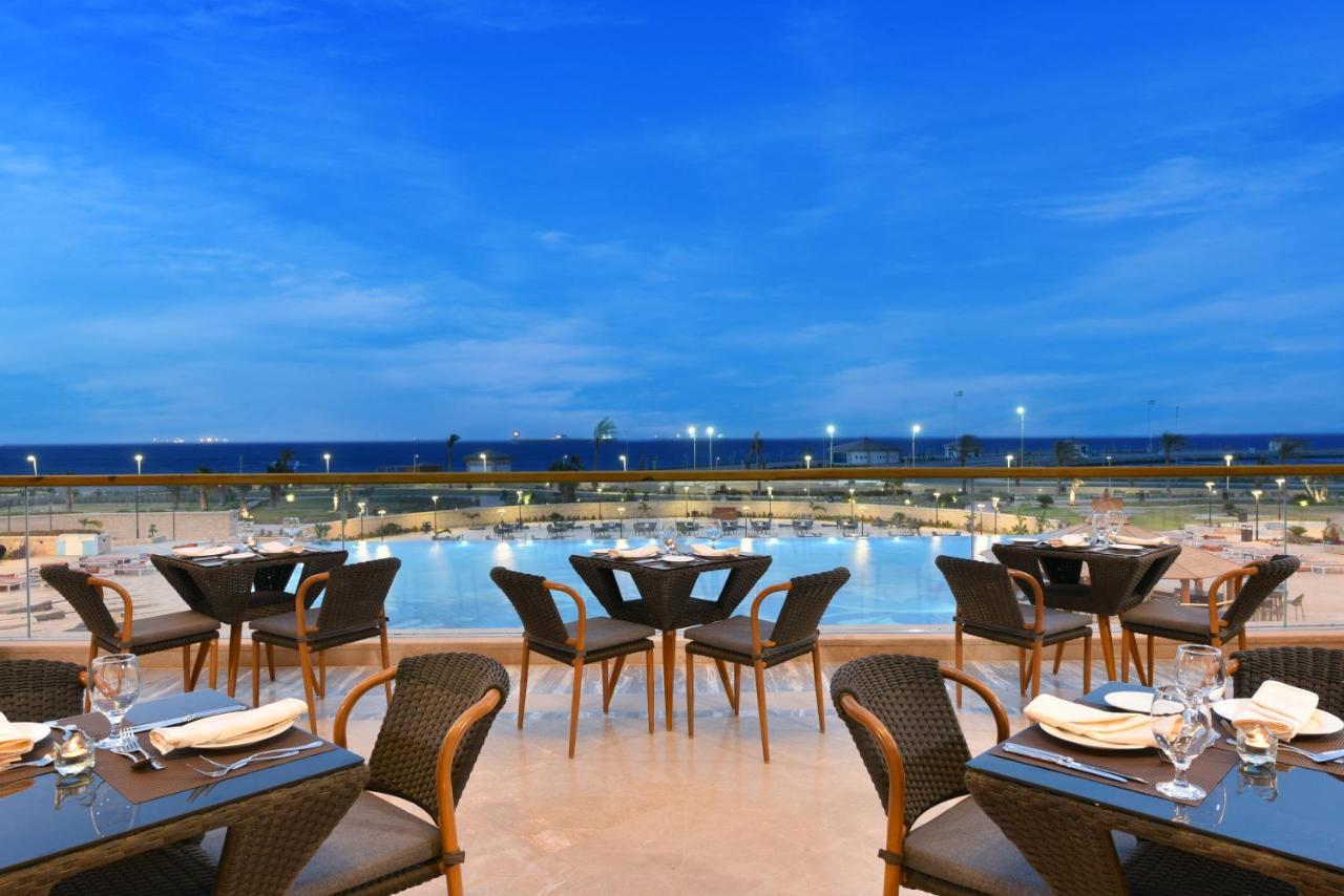 Tolip Resort El Galala Majestic Ain Sukhna Bagian luar foto The photo shows an outdoor dining area with several neatly arranged tables and chairs. The tables are elegantly set with cutlery and plates, suggesting a fine dining experience. In the background, there is a large swimming pool that reflects the blue